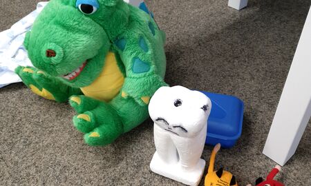 Auf einem Teppich steht ein trauriger Zahn mit schwarzen Karieslöchern in einer Reihe neben einem Plüschdrachen und kleinen Kariesmonstern aus Plüsch.