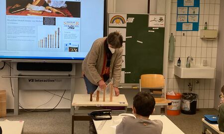 Die Musiklehrerin zeigt vorne auf einem Tisch verschieden große und verschieden farbige Flöten. Ein Schüler sitzt direkt vor ihr an seinem Tisch und schaut aufmerksam zu.