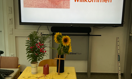 Altar mit Kerze, Blumen und rotem Faden. Im Hintergrund am Smartboard ein Begrüßungsbild mit den Worten "Herzlich Willkommen"
