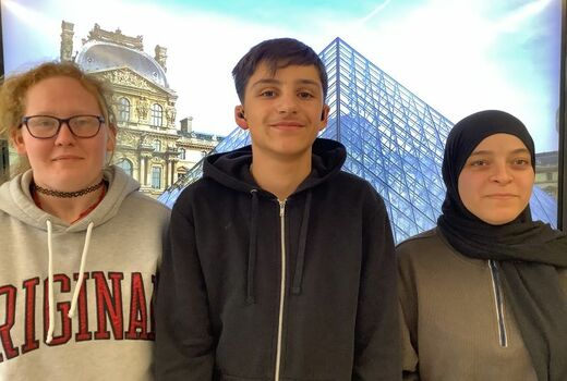 Drei Schüler*innen lächeln den Betrachter an. In der Mitte steht ein Junge, rechts und links ein Mädchen.