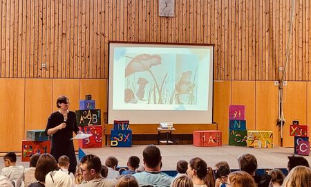 Die Abteilungsleiterin steht vor der dekorierten Bühne und liest den Kindern ein Buch über Mut vor. Die Bilder aus dem Buch werden hinten auf der Leinwand gezeigt.