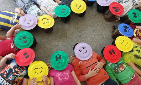 Sechzehn Kinder liegen im Kreis und tragen bunte Masken aus Papptellern, die verschiedene Gefühle in Form von Smileys darstellen.