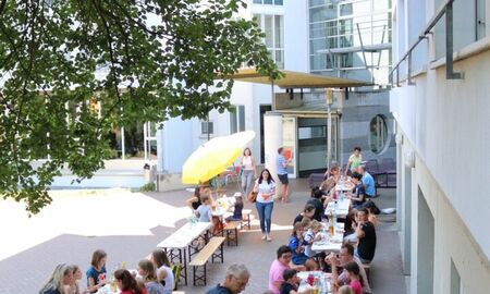 viele Menschen sitzen vor einem Gebäude im Schatten an Biergarnituren und essen und trinken.