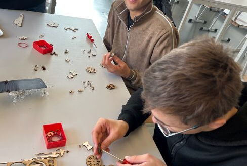Zwei Schüler mit Werkzeug und vielen Kleinteilen an einem Tisch, einer schaut in die Kamera.
