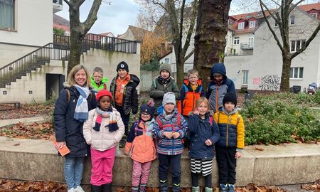 Zehn Schülerinnen und Schüler einer ersten Klasse posieren mit ihrer Lehrerin vor einem Baum in der Stadt für ein Gruppenfoto.
