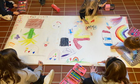 Fünf Schülerinnen sitzen um ein großes Plakat herum und malen es gemeinsam mit Buntstiften an. Auf dem Bild sieht man Regenbögen, eine Blume, eine Sonne, Spielekonsolen und weitere Verzierungen.