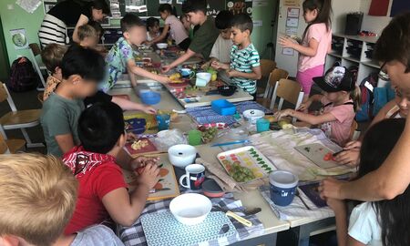 Die Erstklässler und Erstklässlerinnen bereiten an einem langen Tisch gemeinsam das Müsli vor, indem sie unter anderem Obst schneiden.