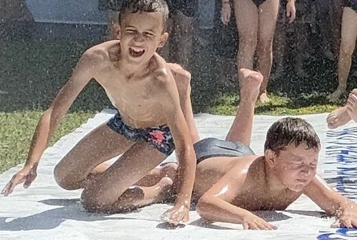 Auf dem Rasen liegt eine LKW-Plane. Zwei Kinder rutschen die nasse Plane herunter. Hinter ihnen warten weitere Kinder in Badekleidung.