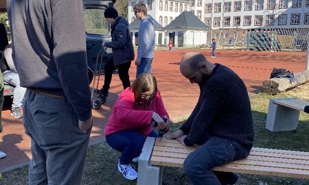 Eine junge Frau schraubt die Sitzfläche auf eine Bank. Ein Lehrer hilft ihr dabei