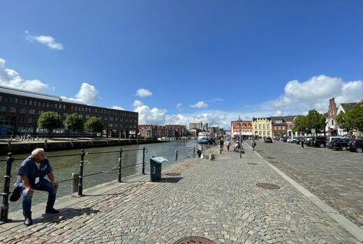 Hafengelände. Links ein Kanal, in der Mitte und rechts gepflasterte Wege. Im Hintergrund un an den Seiten sieht man Häuser, teils in typisch norddeutscher Bauweise.