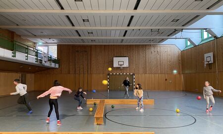 Kinder werfen Bälle über eine Bank in einer Turnhalle