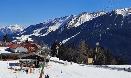verschneite Gebirgslandschaft mit Skipiste, Schlepplift und Gebäuden, entfernt sieht man vier Wintersportler, die mit dem Lift bergauf fahren