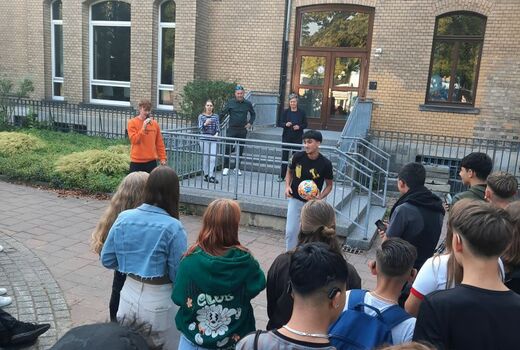 Schüler stehen mit dem Rücken zum Betrachter vor einem Gebäude und beobachten einen Jugendlichen, der einen Ball in der Hand hält. Ein junger Mann mit Mikrofon spricht.