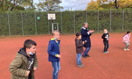 Die Musiklehrerin tanzt und klatscht in einem Halbkreis mit vier Zweitklässlern und einer Zweitklässlerin auf dem Sportplatz.