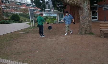 Zwei Schüler sammeln Müll auf dem Schulgelände ein. Ein Junge wirft mit einer Zange Müll in den Eimer. Der Eimer wird von einem anderen Jungen gehalten. Die beiden stehen neben dem Mammutbaum.