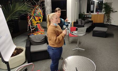 Die Schülerin steht vor der Kamera und hat das Mirko in der Hand. Frau Edelmann steht im Hintergrund und dolmetscht.