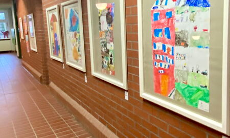 Mehrere Bilderrahmen mit verschiedenen Kunstwerken von Schülerinnen und Schülern hängen in einer Reihe an der Wand. Die sichtbaren Kunstwerke stellen bunte Häuser dar.