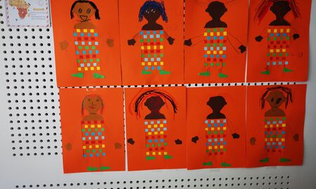 An einer Wand hängen Bilder von Personen, deren Kleidung mithilfe von bunten Papierstreifen und Schlitzen in das Papier gewebt wurde.