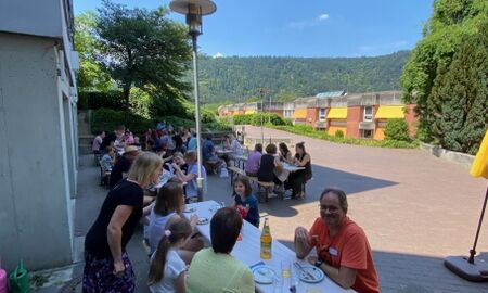 viele Menschen sitzen vor einem Gebäude im Schatten an Biergarnituren und essen und trinken.
