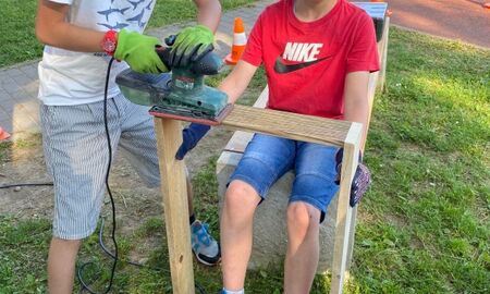 Ein Schüler hebt einen Holzrahmen fest. Ein anderes Kind schleift diesen mit einer Schleifmaschine