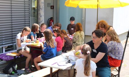 Kinder und zwei Lehrerinnen sitzen zusammen unter einem Sonnenschirm und basteln