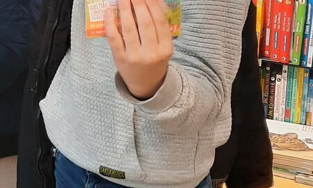 Ein Schüler steht in der Buchhandlung vor einem Bücherregal und hält das Buch "Mission Roboter" in die Kamera.