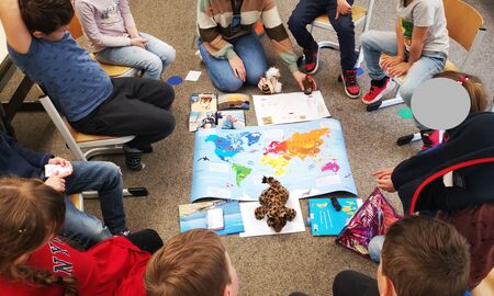 Neun Erstklässler und Erstklässlerinnen sitzen auf Stühlen im Sitzkreis. Die Lehrerin sitzt vorne auf dem Boden. In der Mitte liegen eine Weltkarte und verschiedene persönliche Mitbringsel der Kinder.