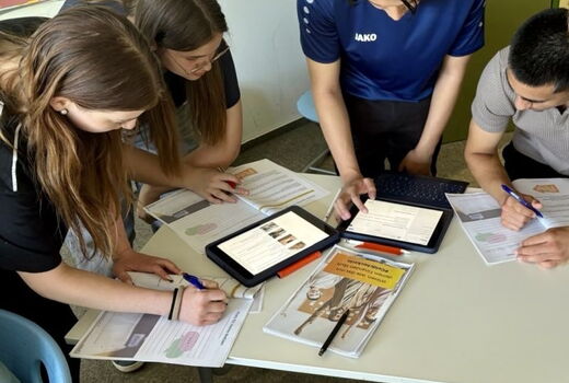 Zwei Jungs und zwei Mädchen arbeiten an einem Tisch. Vor ihnen liegen Arbeitshefte und zwei Tablets.