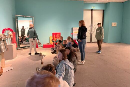 Zwölf Schüler und eine Lehrerin sitzen bzw. stehen in einem Raum, vor sich verschiedene Gegenstände bzw. Bekleidung aus dem Leben römischer Soldaten. Eine Frau vor ihnen erklärt. Sie hält einen Helm in der Hand.