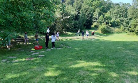 Schülerinnen und Schüler stehen oder laufen auf einer großen weitläufigen Wiese im Schatten vieler Bäume