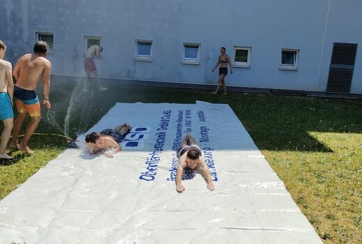 Auf dem Rasen liegt eine LKW-Plane. Zwei Kinder rutschen nebeneinander die nasse Plane herunter.