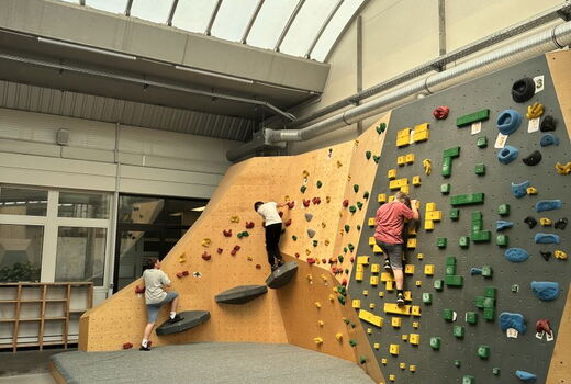 Blick in eine Boulderhalle. Rechts an einer Wand klettern drei Kinder.