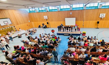 Die Einschulungsfeier ist von oben zu sehen. Die neuen Erstklässler und Erstklässlerinnen sitzen in den ersten Reihen. Dahinter sitzen ihre Eltern. Vorne auf der Bühne stehen Schüler und Schülerinnen.