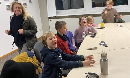 Eine Lehrerin läuft um einen Tisch herum, an dem sechs Kinder sitzen. Diese lachen