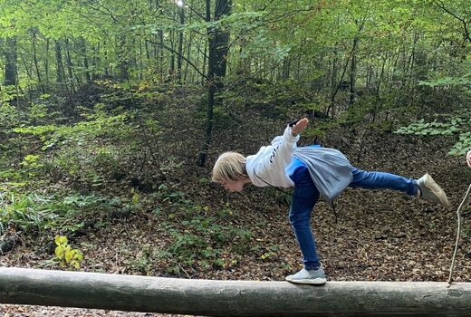 Kind balanciert auf einem Baumstamm. Auf dem Weg in den Wald gibt es Abenteuer zu bestehen.