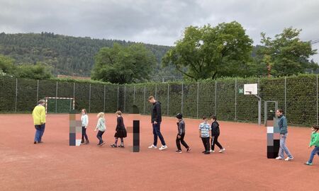 Die Musiklehrerin läuft vorne, ihr folgen zehn Kinder und zwei Erwachsenen. Alle laufen in einer Reihe hintereinander über den Sportplatz.