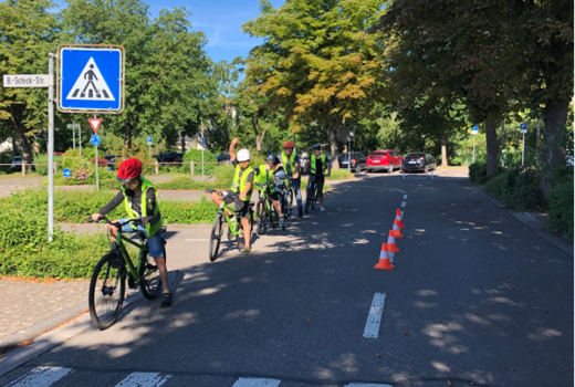 Sechs Kinder in Warnwesten und Helmen warten auf Fahrrädern auf einer Übungsstraße in einer Reihe hintereinander an einem Zebrastreifen.