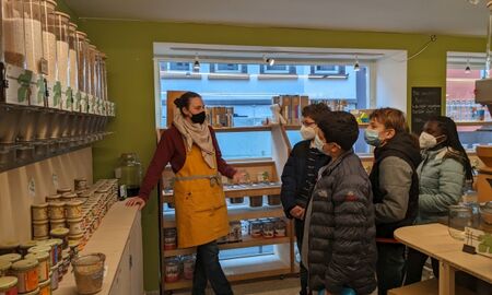 Vier Kinder und eine Frau im Unverpacktladen, die Frau scheint zu erklären, im Hintergrund ist das Schaufenster