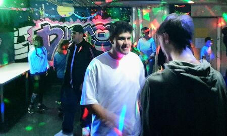 Zwei Jungen stehen sich auf der Tanzfläche gegenüber. Einer von ihnen lächelt. Im Hintergrund sind andere SchülerInnen zu sehen. Die Lichtanlage leuchtet.