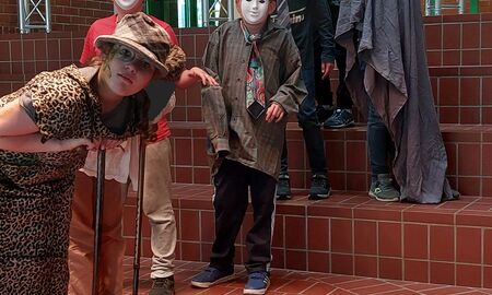 Eine verkleidete Schülerin steht vorne und schaut in die Kamera. Hinter ihr auf Stufen stehen fünf weitere Kinder mit weißen Masken im Gesicht.