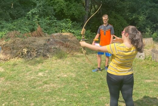 Im Vordergrund steht ein Mädchen mit einem Bogen in Rückenansicht. Sie schießt gerade einen Pfeil ab. Im Hintergrund steht ein junger Mann und überwacht ihren Schuss.