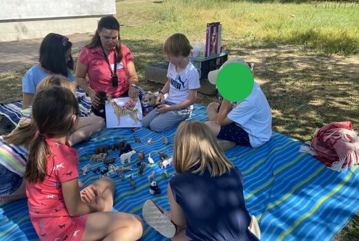Eine Lehrerin zeigt den Kindern ein stück Papier mit einem Tier darauf. Auf einem Teppich in der Mitte liegen viele Schleichtiere. Die Kinder sitzen aufmerksam drum herum und Hören zu