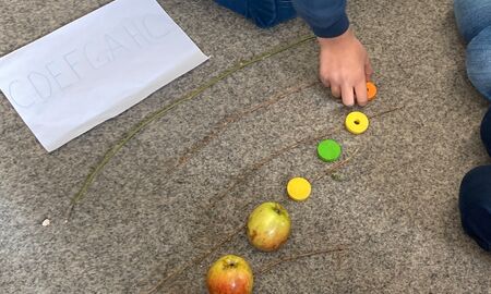 Auf dem Teppichboden liegen die Äste als Notenlinien. Auf und zwischen den Linien liegen die Äpfel und Holzkreise als Notenköpfe in einer Tonleiterabfolge.