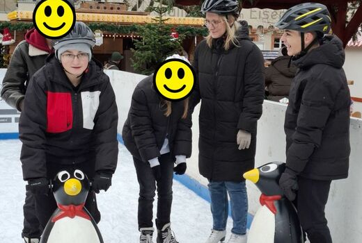 Vier Schüler und eine Lehrerin stehen auf der Eisbahn. Zwei Kinder heben sich an einer Stützhilfe in Form eines Pinguins fest.