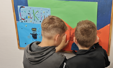 Zwei Jungs stehen vor einer Pinnwand und pinnen ein orangenes Bild an. Die Pinnwand hat einen blauen Hintergrund und trägt den Schriftzug "Cool Kids"