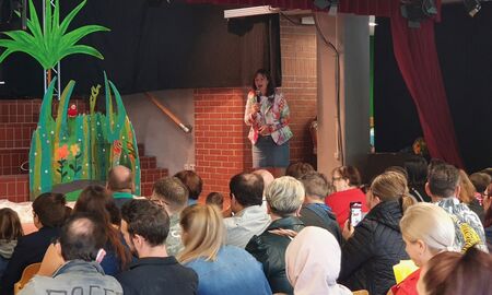 Die Schulleitung spricht mit einem Mikrofon auf einer Bühne vor den Eltern und Erziehungsberechtigten der neuen Erstklässler und Erstklässlerinnen.