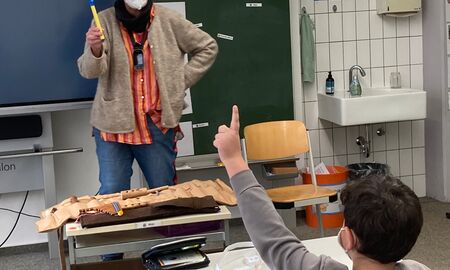 Die Musiklehrerin hält eine Flöte hoch. Vor ihr auf dem Tisch liegen noch weitere Flöten in einem Ledereinband. Ein Schüler sitzt an seinem Tisch vor ihr und meldet sich.