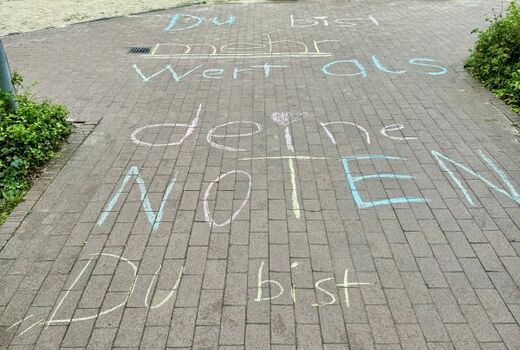 Auf einem gepflasterten Weg auf dem Schulhof steht in großen Buchstaben mit bunter Kreide geschrieben: Du bist mehr wert als deine Noten. Du bist wertvoll.
