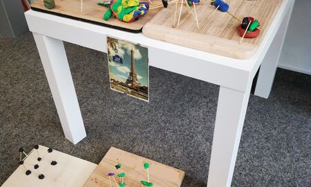 Auf und vor einem kleinen weißen Tisch stehen auf Holzbrettchen zahlreiche unterschiedliche Nachbauten des Eifelturms aus Zahnstochern und Knete. An dem Tisch hängt ein Bild des Eifelturms.