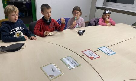 Zwei Mädchen und zwei Jungen sitzen an einem Tisch. Auf der Tischmitte liegen Karten. Ein Mädchen spricht in ein Mikrofon